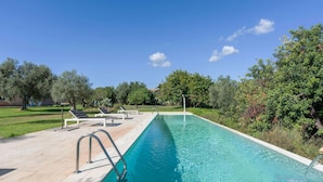 Une piscine rectangulaire à l’eau d’un bleu limpide, entourée d’une terrasse carrelée. On y trouve des chaises longues et un escalier en métal qui mène à l’intérieur de la piscine.