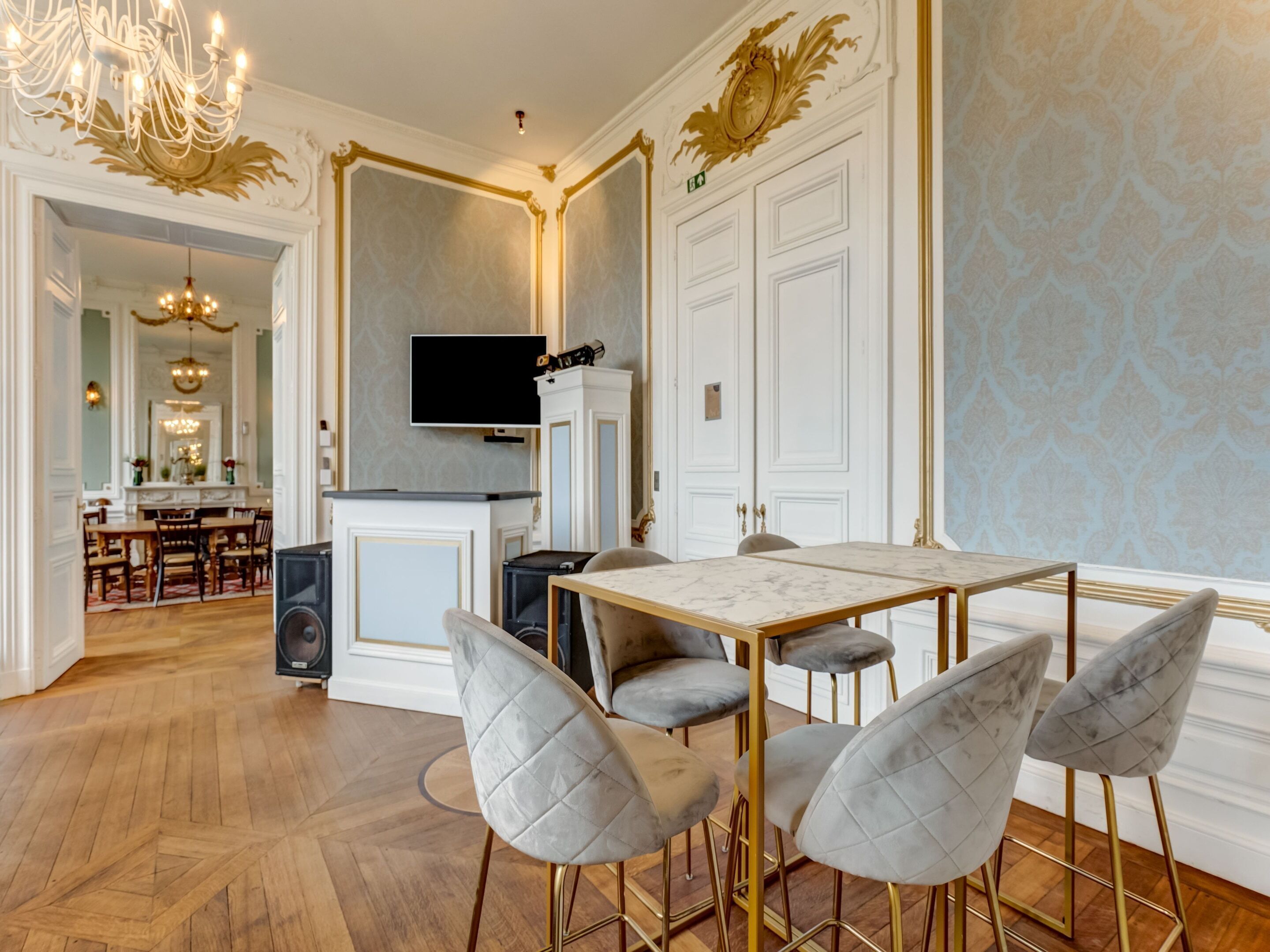 A dining area with a marble table, four grey upholstered chairs, a chandelier, and a television above a cabinet.