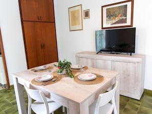 A dining area with a wooden table set for four, surrounded by white chairs. A television is mounted on a cabinet above the table. - Vue Merveilleuse Apartment (Castellammare del Golfo)