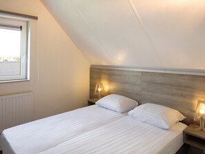 Ein Schlafzimmer mit geneigter Decke, einem Fenster mit Jalousien, einem Bett mit weißer Bettwäsche, zwei Nachttischen mit Lampen und einem Heizkörper.