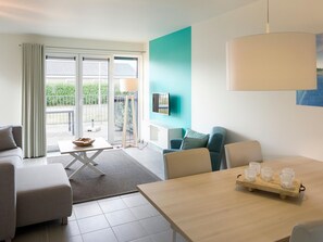 Ein modernes Wohnzimmer mit einer Couch, einem Esstisch mit Gläsern, einem Fernseher und Blick ins Freie.
