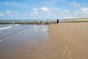Ein sandiger Strand mit einem steinernen Wellenbrecher, blauem Himmel und ruhiger See.