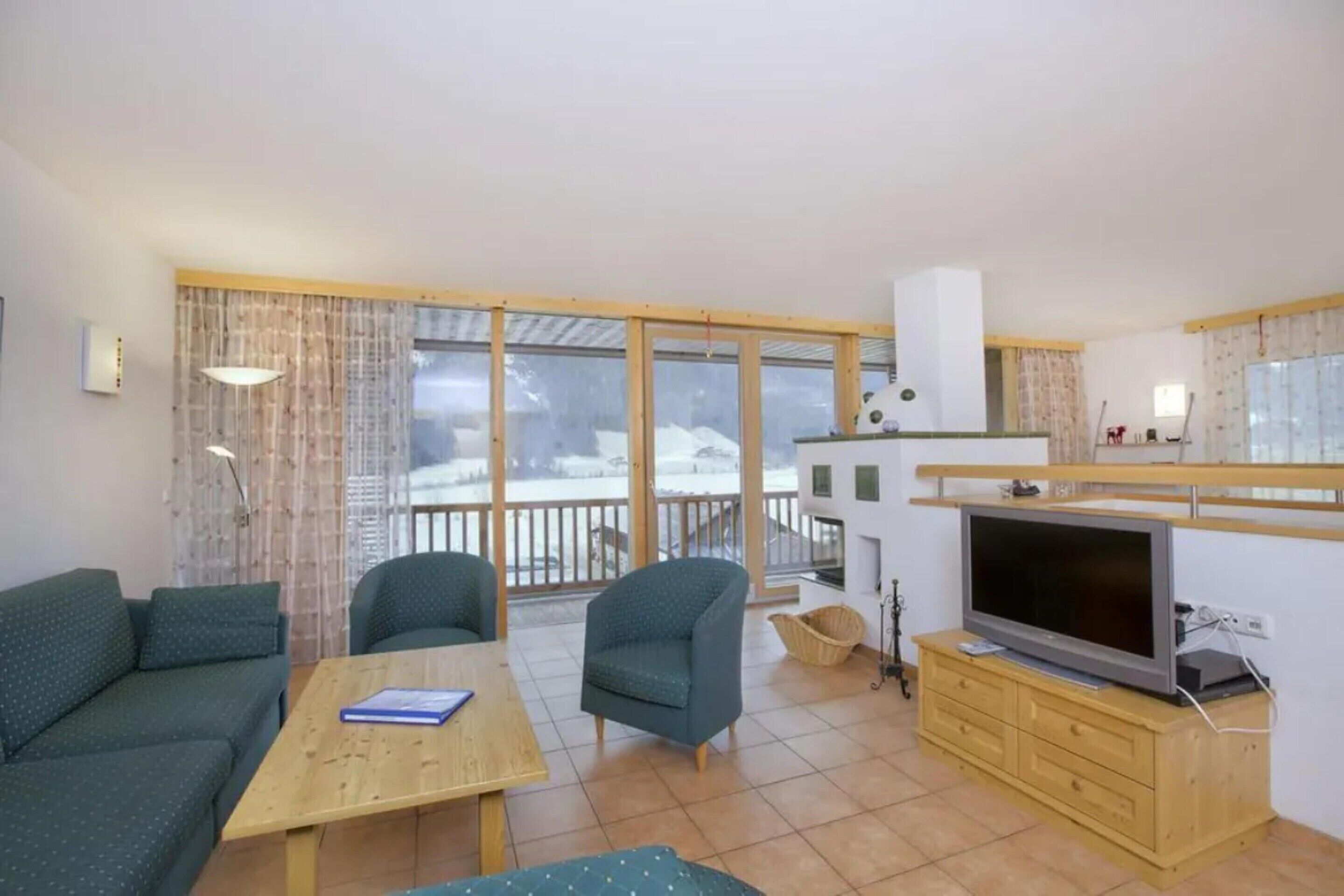 A furnished living room with a sofa, chairs, a table, a television, and a balcony with a view of a snowy landscape.