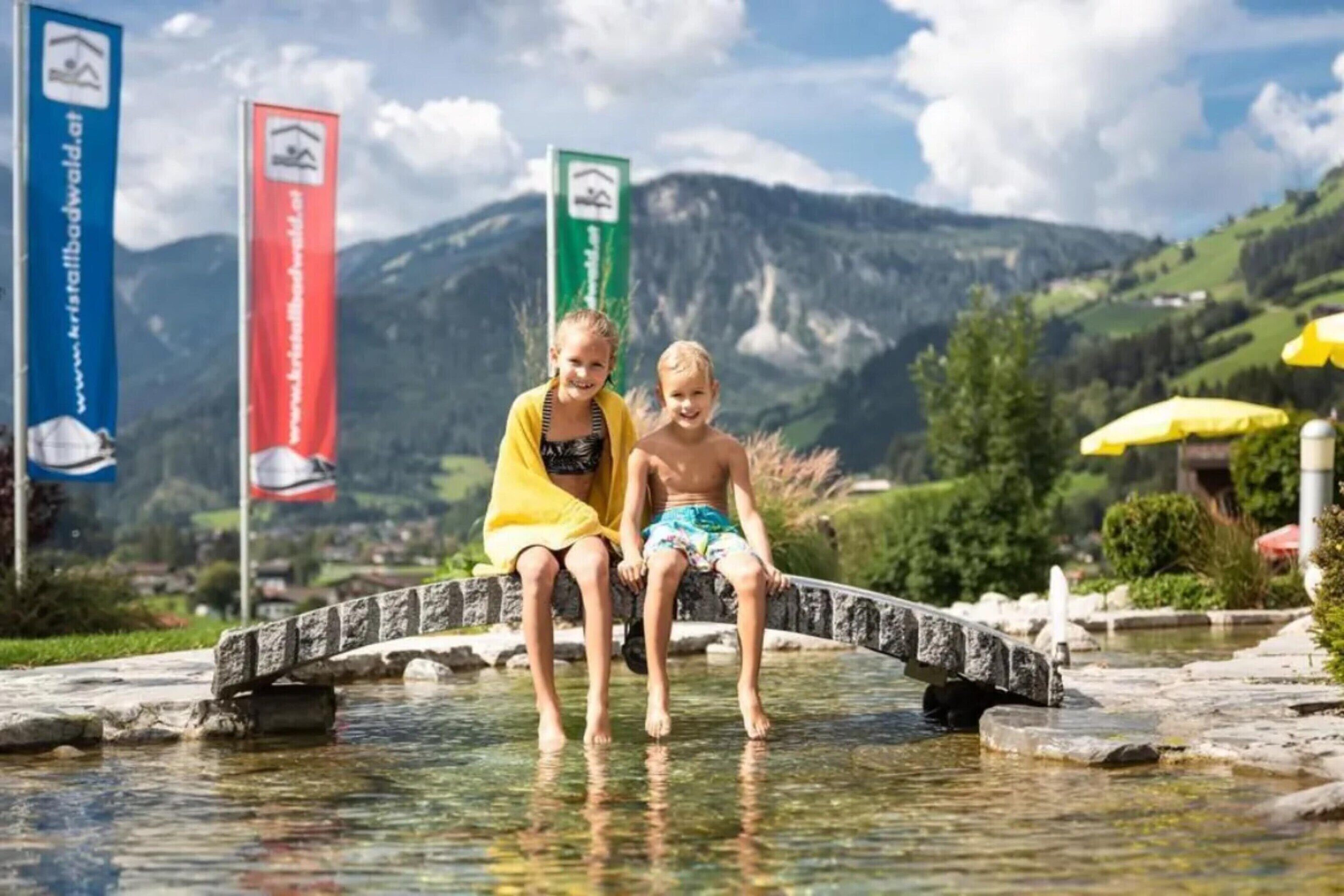 Zwei Personen sitzen auf einer Steinsbrücke über einem klaren Gewässer, im Hintergrund Berge.