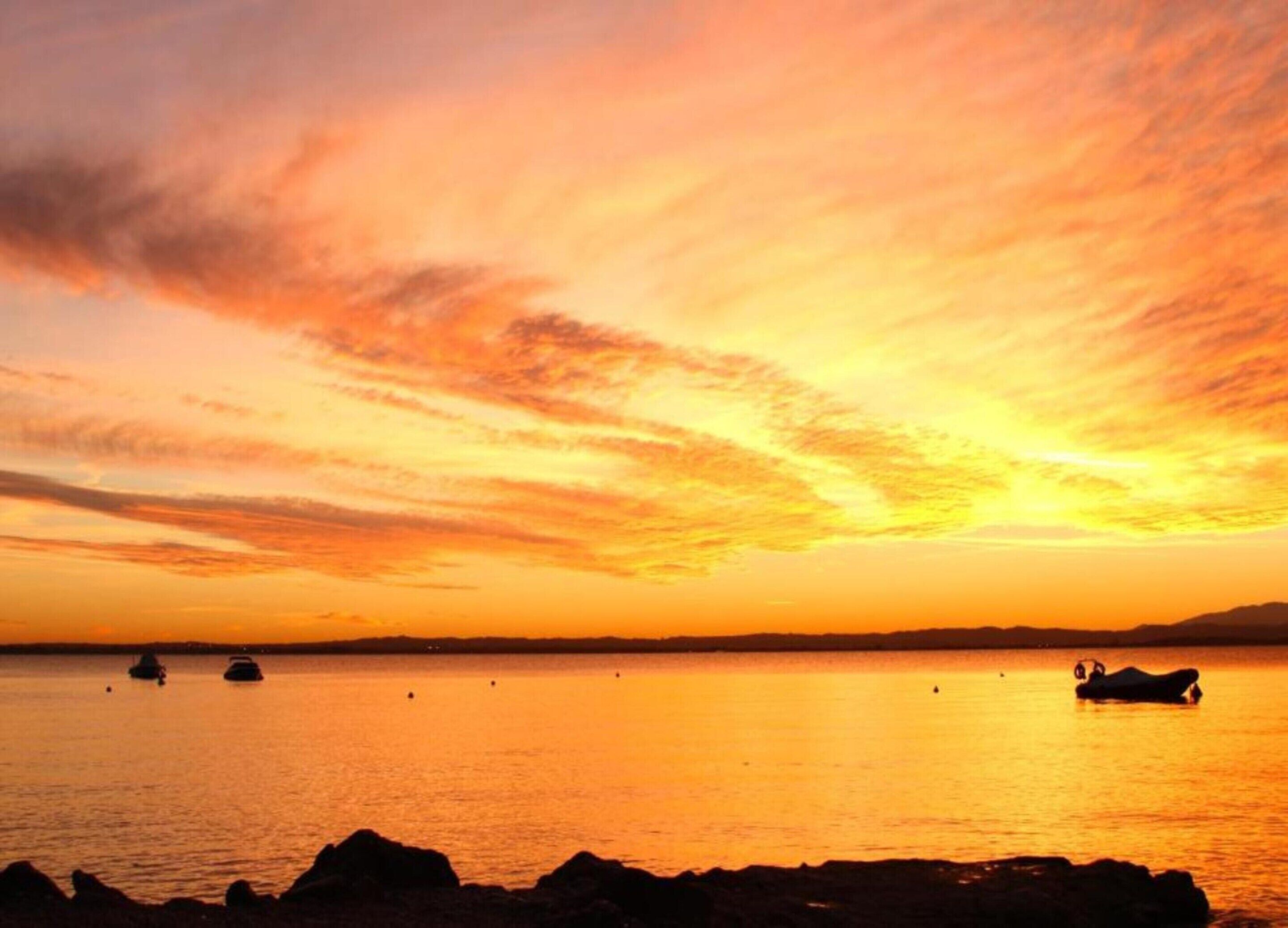 A sunset over a calm body of water with boats and rocks in the foreground.