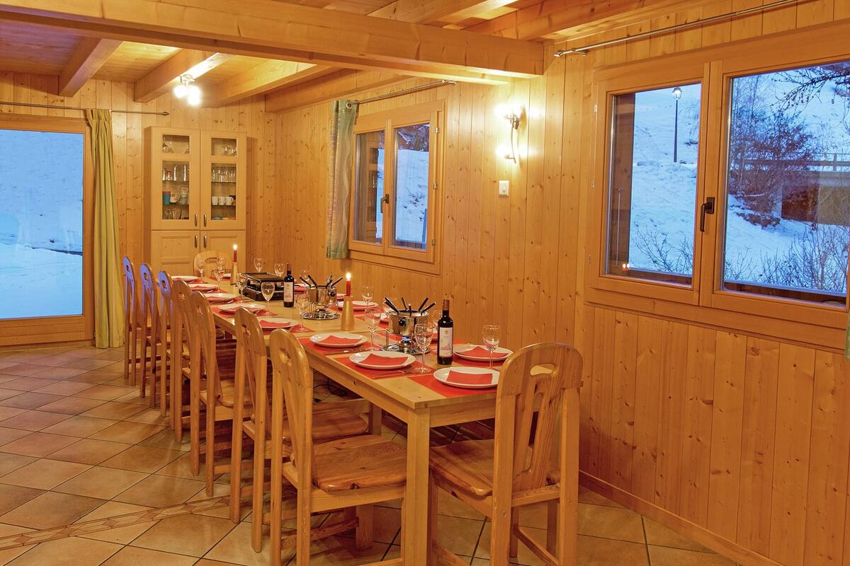 A wooden dining room with a long table set for a meal, wooden chairs, and a view of a snowy landscape through large windows.