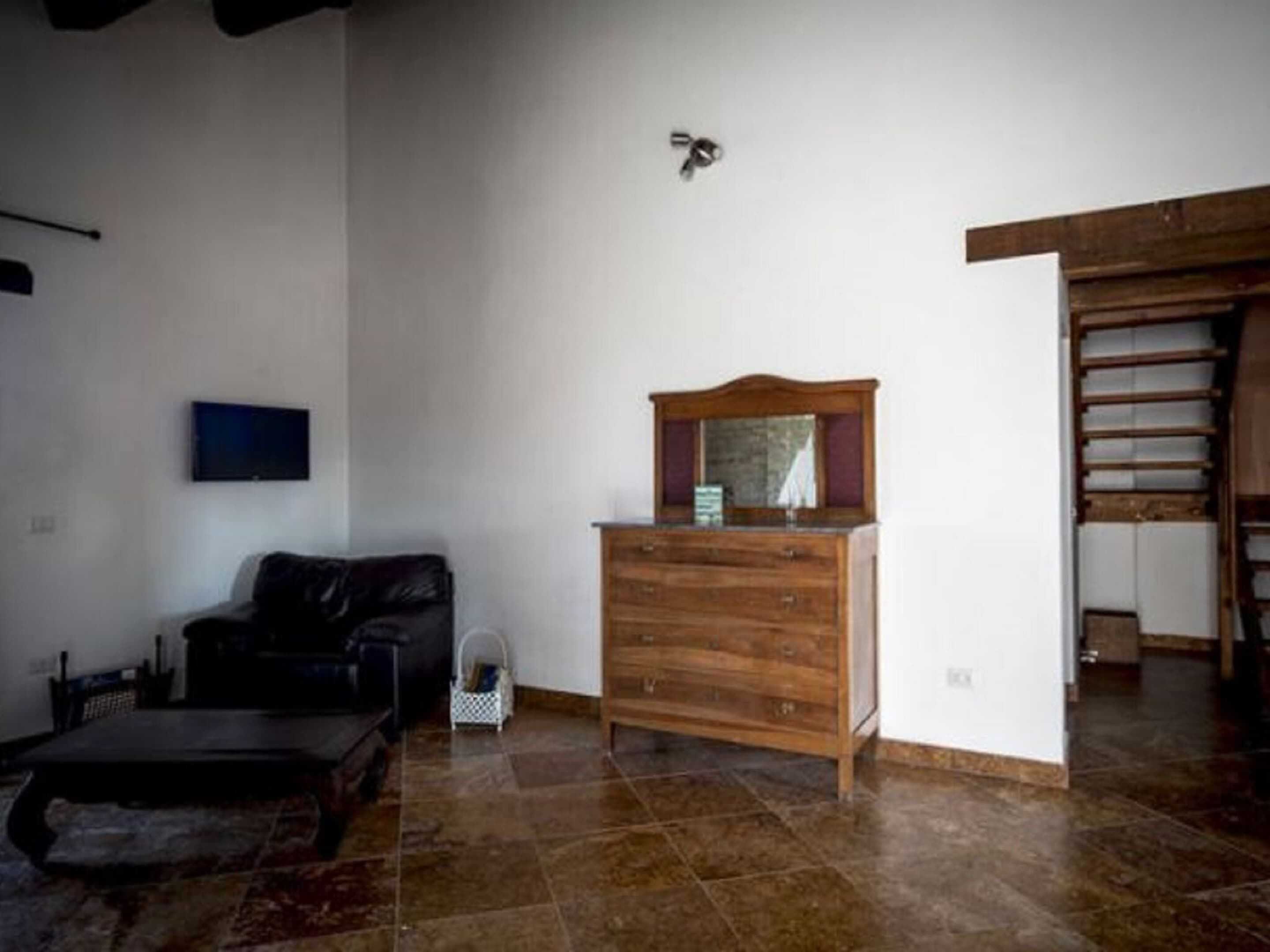 Una habitación con un aparador de madera, un televisor montado en la pared, un sofá de cuero negro y suelo de baldosas.