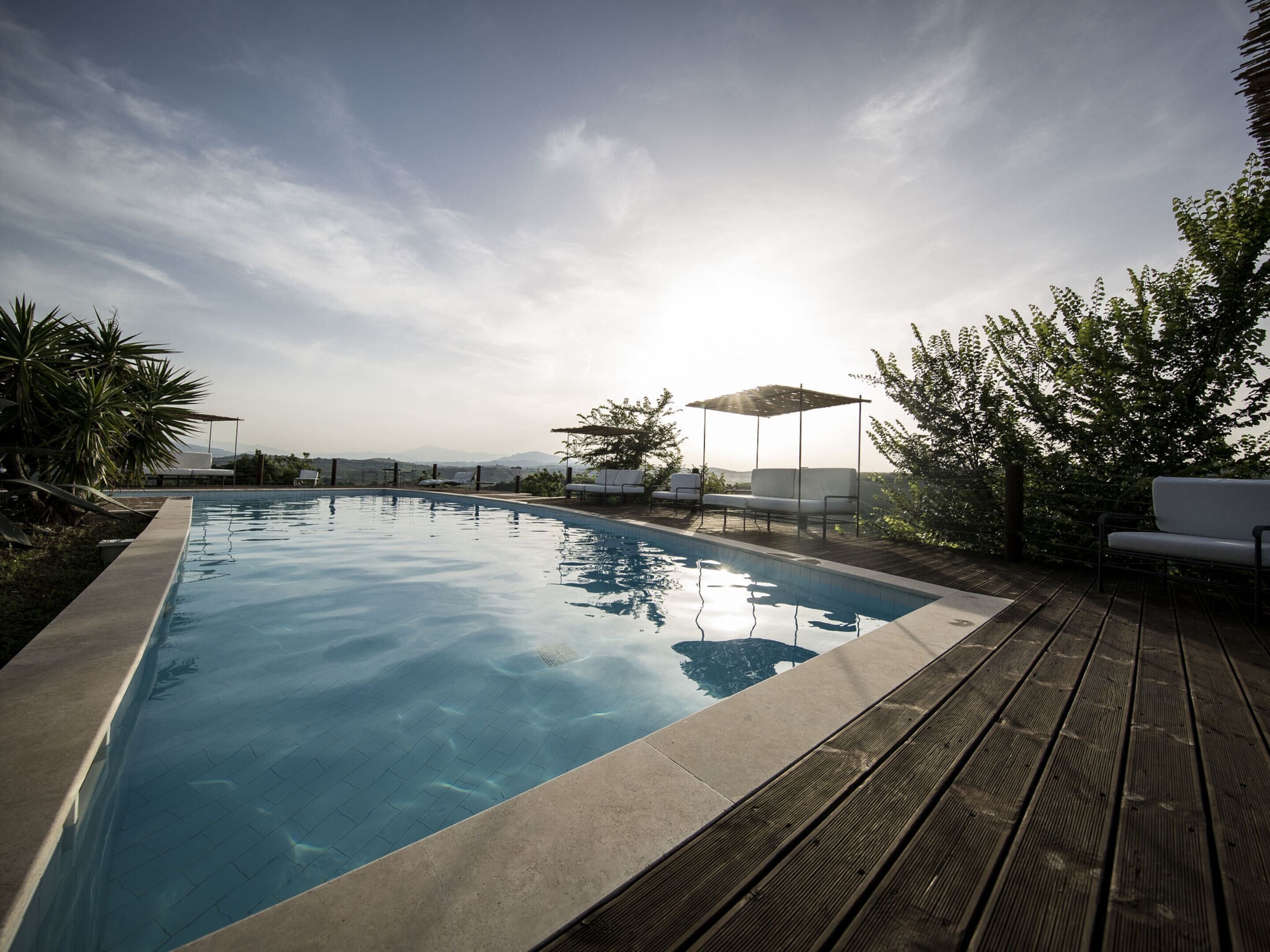 A swimming pool with a wooden deck, surrounded by greenery and a clear sky.