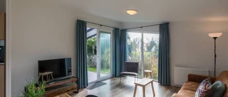 Un salon moderne avec une table à manger en bois, un canapé, un téléviseur et une grande porte-fenêtre coulissante donnant sur un jardin.