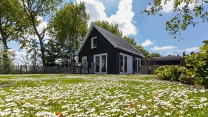 A black house with a white door and windows, surrounded by a grassy area with white flowers. - Holiday Home in Vrouwenpolder With Garden (Vrouwenpolder)