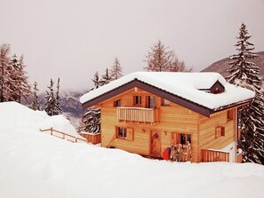 Una cabaña de madera con balcón, rodeada de árboles y montañas cubiertas de nieve.