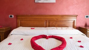 Une chambre d’hôtel avec une tête de lit en bois, une couvre-lit blanche et une décoration en forme de cœur composée de pétales de roses rouges sur le lit.