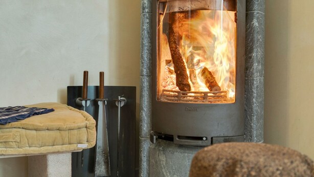 Une cheminée en pierre avec une porte en verre, des bûches qui brûlent à l’intérieur, et un lit avec une couverture.