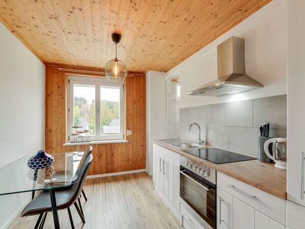 A modern kitchen with wooden ceiling, a dining table with chairs, a large window, and stainless steel appliances.