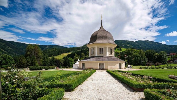 Une église dotée d’un clocher remarquable, entourée de jardins bien entretenus et surplombée par un paysage montagneux.