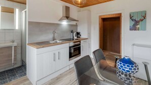 Une cuisine moderne avec des armoires blanches, un plan de travail en bois, un évier en acier inoxydable et une hotte noire. On y trouve un coin repas avec une table et des chaises en verre, ainsi qu’un vase bleu posé sur la table.
