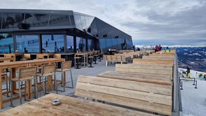 Un espace extérieur aménagé avec des tables et des chaises en bois, un bâtiment moderne doté de grandes fenêtres et une vue sur des montagnes enneigées.