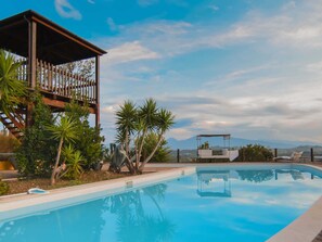 A swimming pool with a wooden deck and lounge chairs, surrounded by tropical plants and a view of mountains.