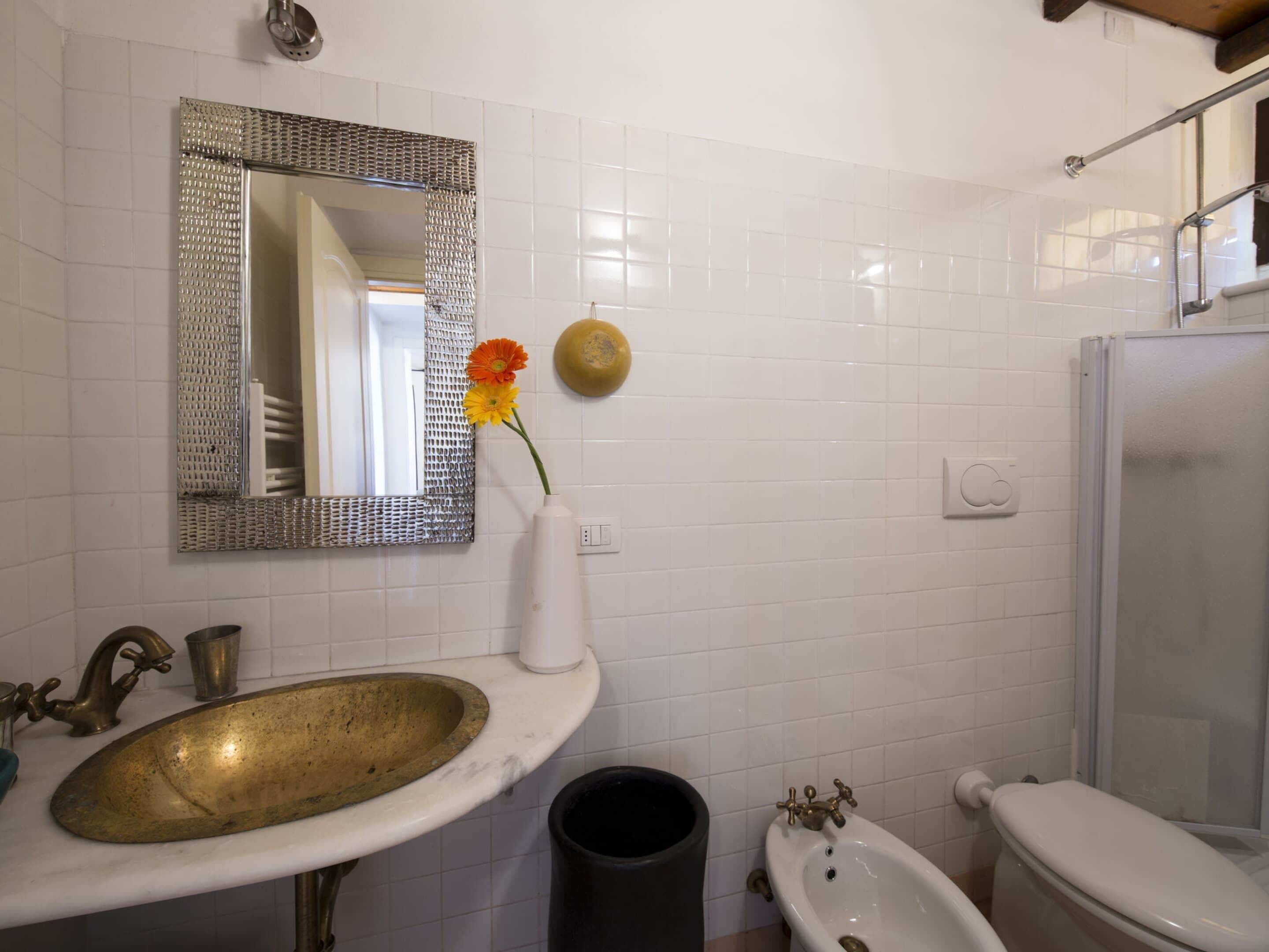 A bathroom with a white tiled wall, a brass sink, a white pedestal sink, a bidet, and a mirror with a decorative frame.