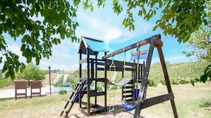 Un parque infantil con una estructura de madera, que incluye un tobogán y columpios, situado en un área cubierta de césped con árboles y cielo despejado.