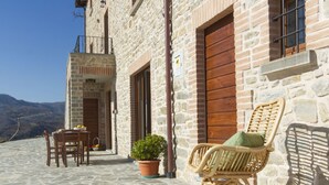 A stone building with a wooden door and window, a wicker chair with a cushion, a table with chairs, and a potted plant.