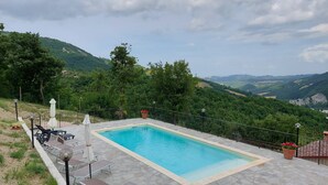 A rectangular pool with a stone deck, surrounded by a metal fence, and overlooking a mountainous landscape.