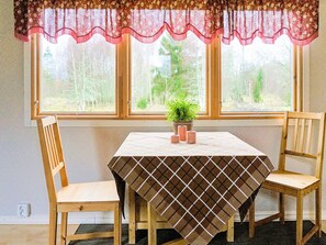 A dining area with a table covered by a checkered tablecloth, two wooden chairs, a potted plant, and a window with a floral valance. - 4 Person Holiday Home in Sandhem (Sandhem)