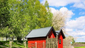 A red wooden building with a sloped roof, surrounded by a wooden fence, set in a grassy area with trees in the background. - 4 Person Holiday Home in Sandhem (Sandhem)