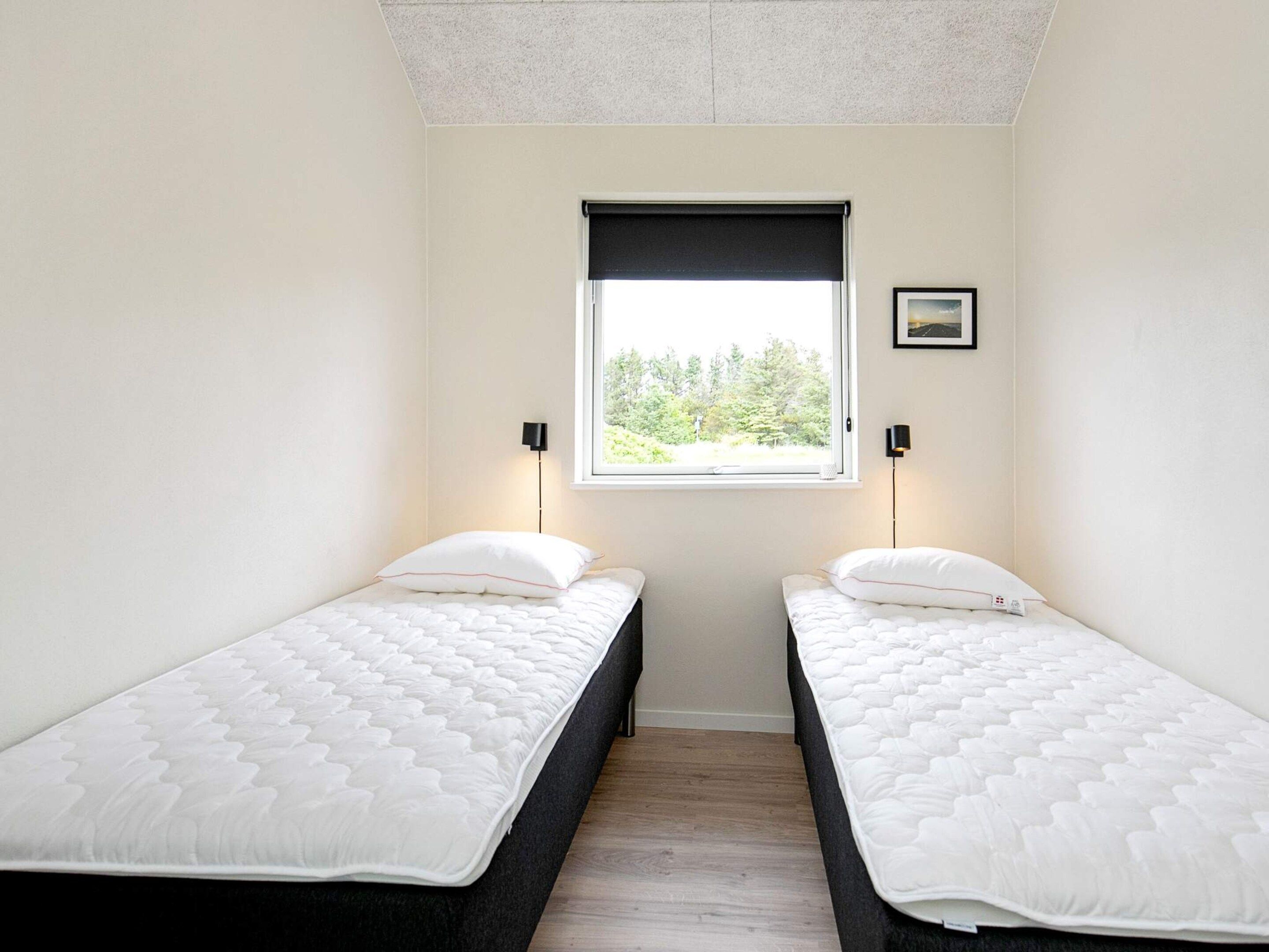 A room with two single beds, a window with a view of greenery, and two wall-mounted lamps.