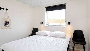 A bedroom with a bed, two chairs, a window with a black blind, a wall-mounted lamp, and a hanging bag.