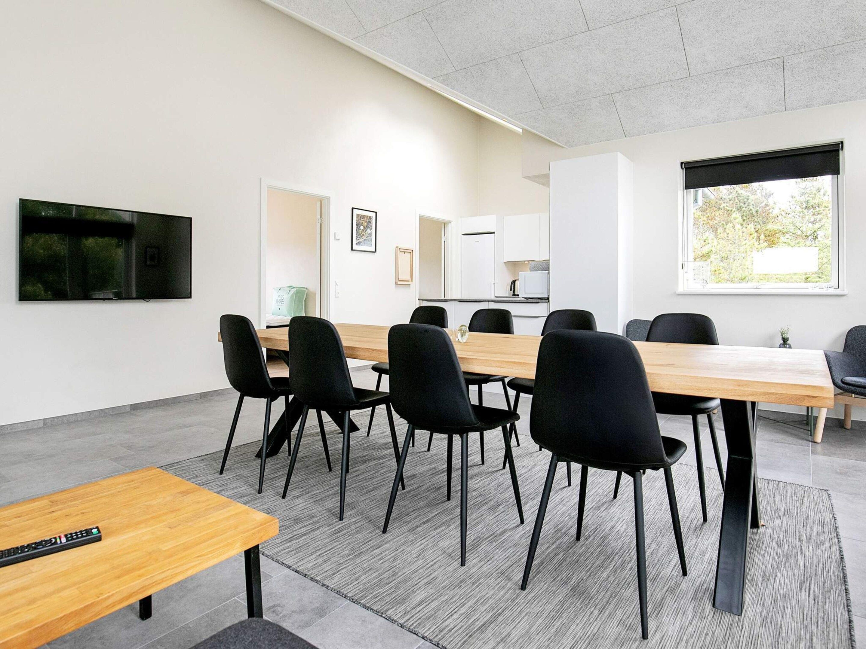 A conference room with a long table, black chairs, a TV on the wall, and a window with blinds.