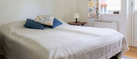 Un dormitorio con cama, almohadas, una mesita de noche con lámpara, una ventana con cortinas y un radiador.