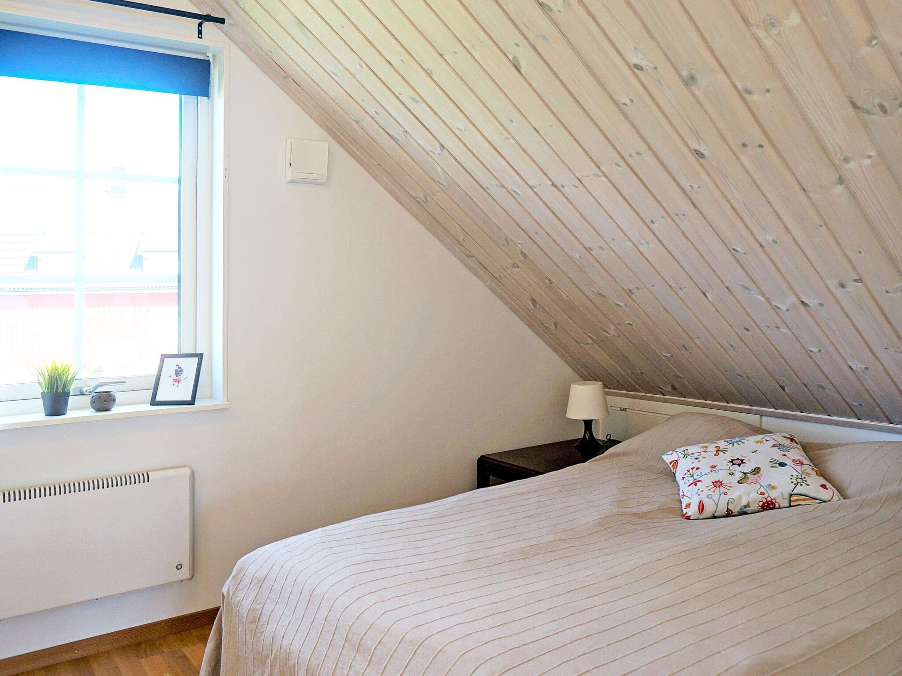 A bedroom with a sloped ceiling, a bed with a floral pillow, a bedside table with a lamp, a radiator, and a window with a plant and picture frame.