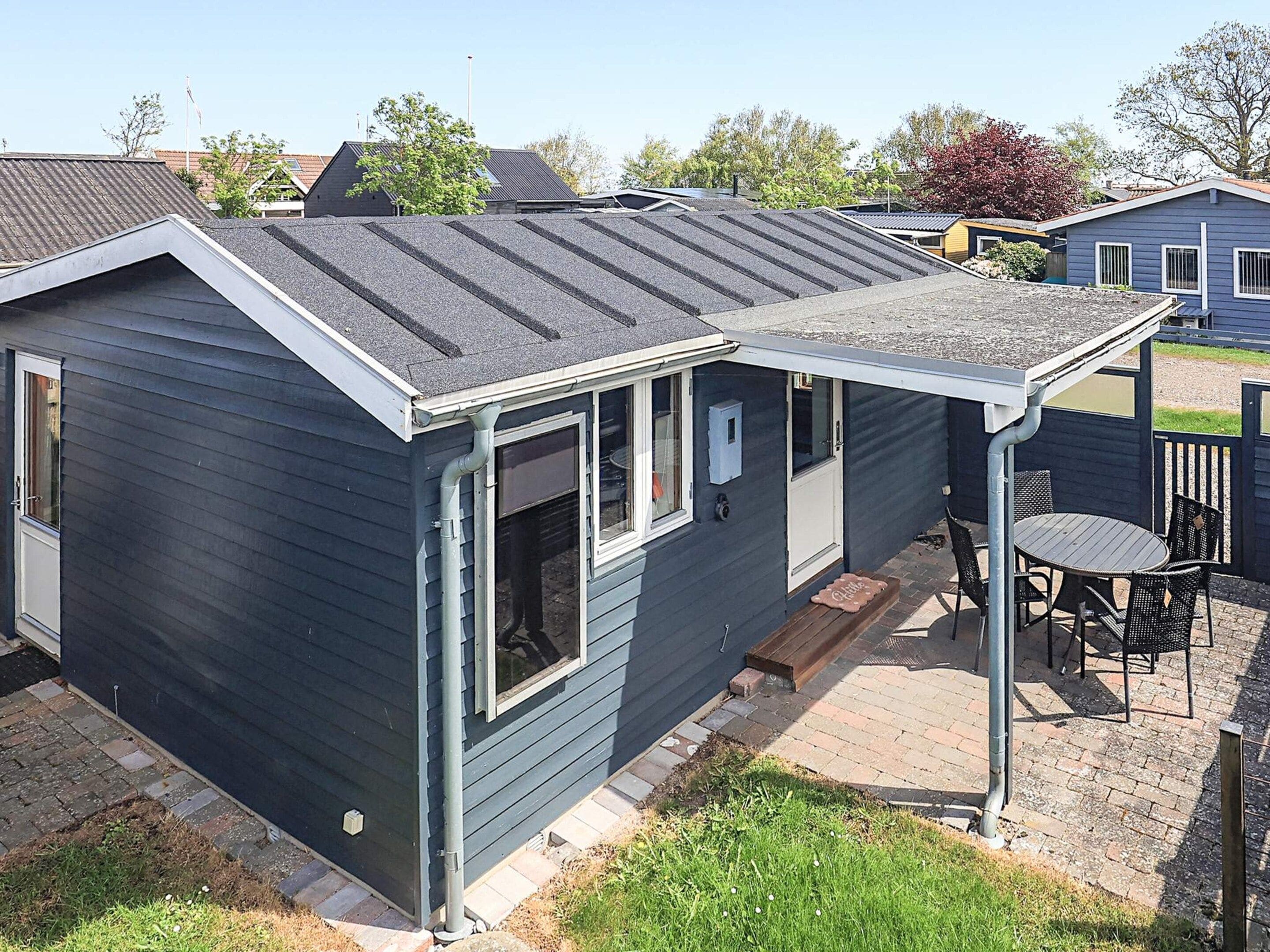A small, single-story house with a dark exterior, a sloped roof, and a covered patio area with a table and chairs.