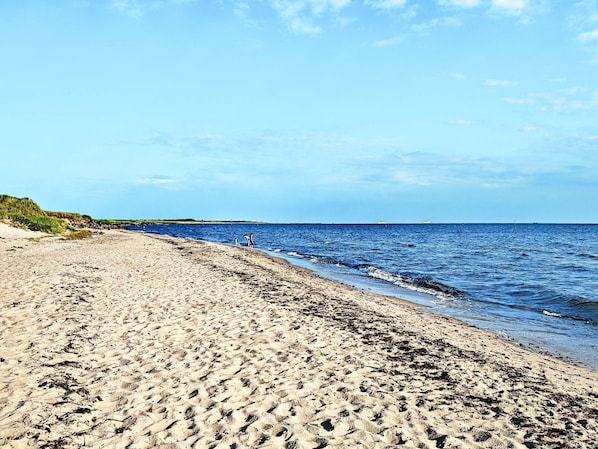 En sandstrand med milda vÄgor, en klar himmel och bÄtar i fjÀrran.
