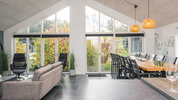 A spacious room with a large dining table, modern chairs, and a sofa. There are large windows providing a view of the outdoors. - Holiday Home in Glesborg (Glesborg)