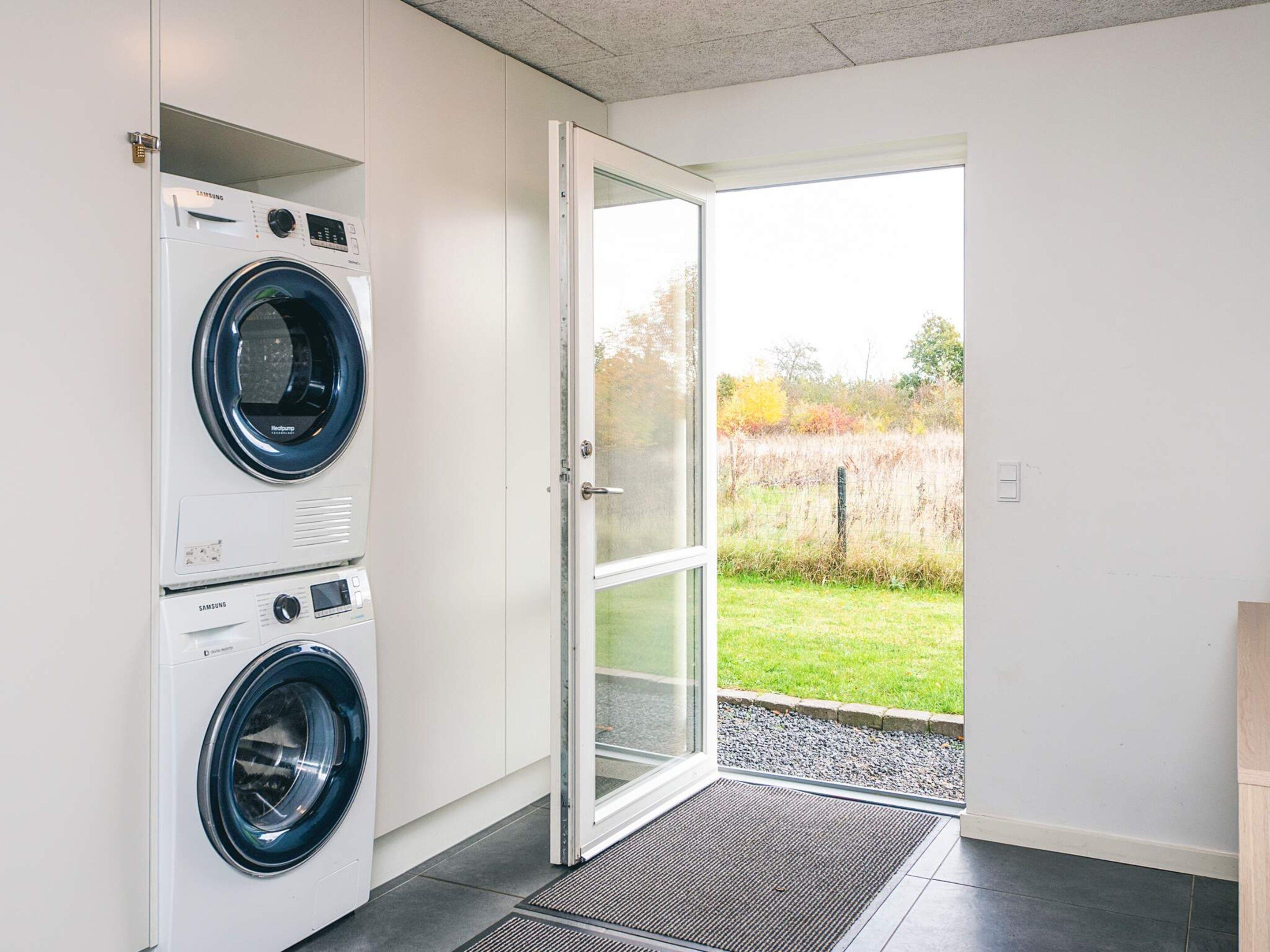 A laundry room with two stacked washing machines, a glass door leading to an outdoor area with a grassy field and trees.