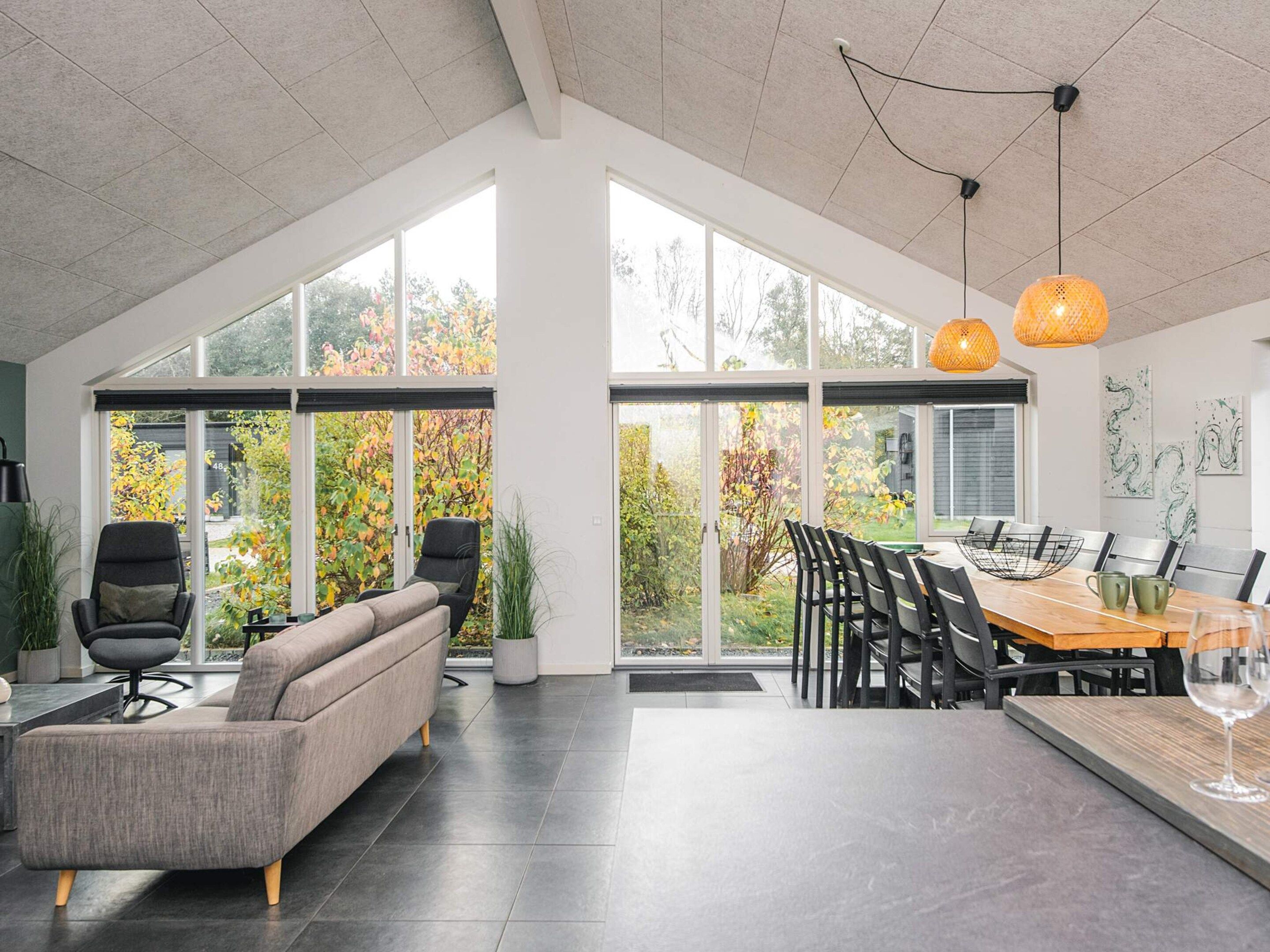 A spacious room with a large dining table, modern chairs, a sofa, and large windows offering a view of a garden.