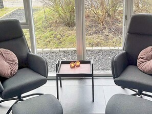 A modern room with two grey armchairs, a small table with two candles, and a view of a garden through large windows.