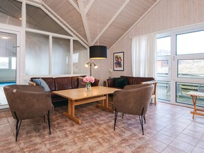 Un amplio espacio habitable con techo de madera, ventanas grandes, un sofá de cuero marrón, dos sillones, una mesa de centro de madera y una lámpara de pie.