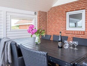 Un patio techado con pared de ladrillo, una mesa con sillas, un florero con flores y una botella de champán.