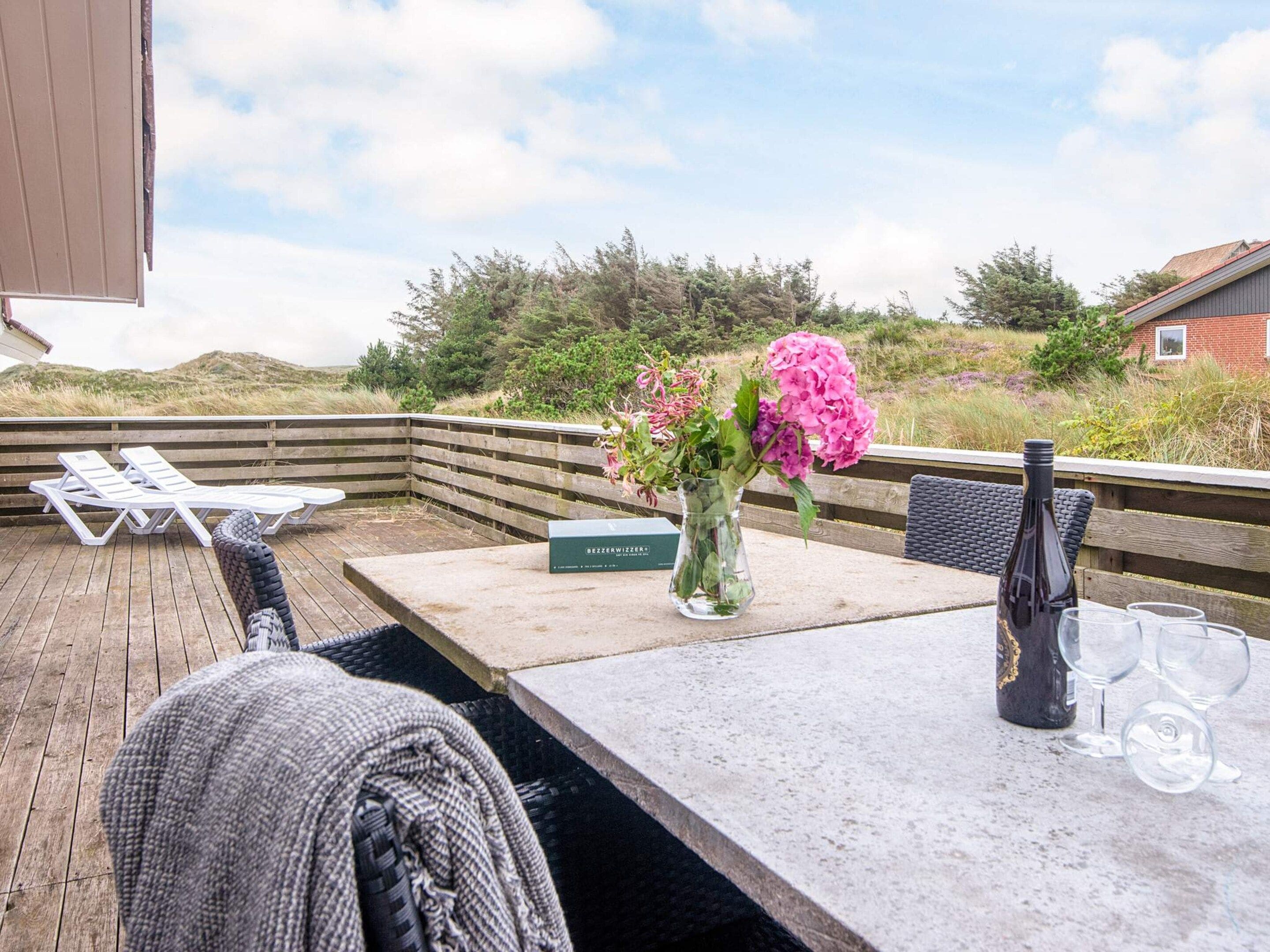 Eine Holzterrasse mit Tisch, Stühlen, einer Blumenvase und einer Weinflasche.
