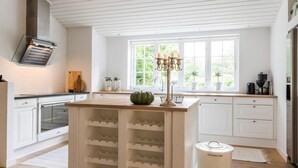 A kitchen with white cabinetry, a central island with wine racks, a window with a view of greenery, and a built-in oven. - 5 Star Holiday Home in Henne (Henne)