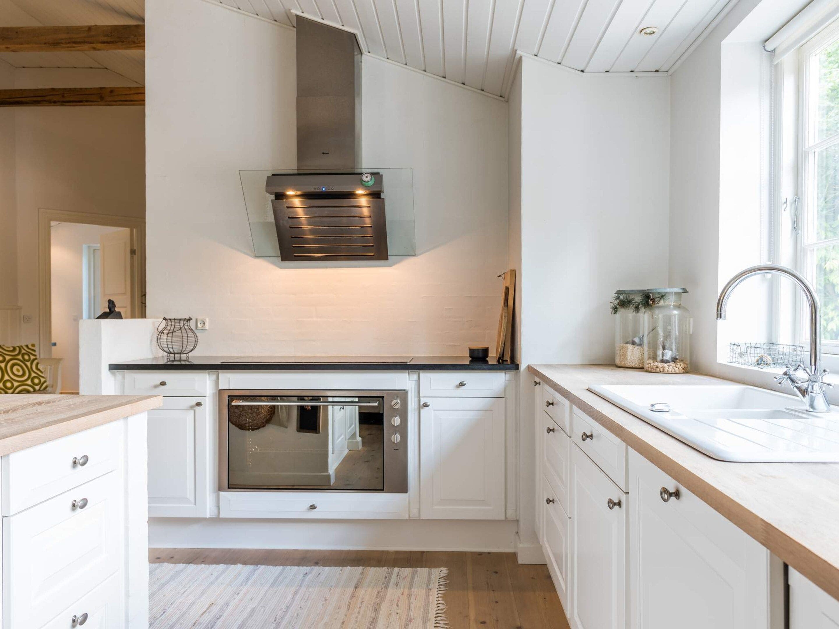 A modern kitchen with white cabinetry, a stainless steel oven, a built-in range hood, and a window providing natural light.