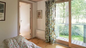 Ein Schlafzimmer mit Bett, Tür, Fenster mit Vorhängen und Blick ins Freie.