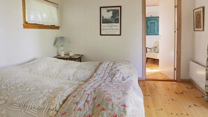 Ein Schlafzimmer mit Holzboden, einem Bett mit blumigem Überwurf, einem Nachttisch mit Lampe, einem gerahmten Bild an der Wand und einer Tür zu einem anderen Raum.