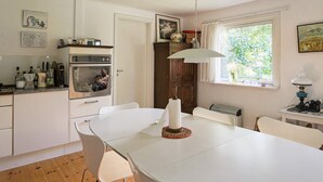 Eine Küche mit weißen Schränken, einem Esstisch, Stühlen, einem Backofen, einem Fenster mit Vorhängen und einer Pendelleuchte.