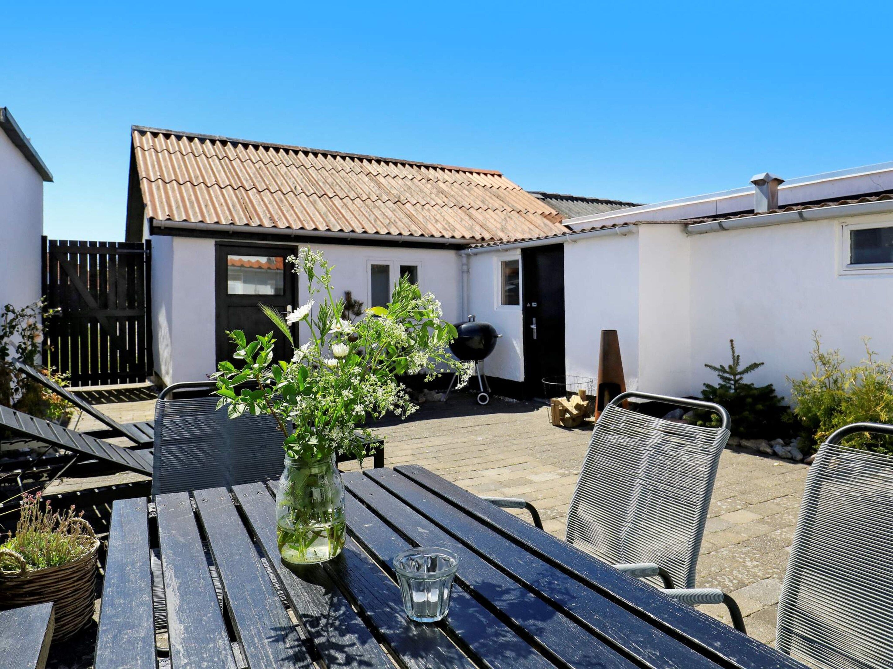 Un espace patio comprenant une table, des chaises, un barbecue et un bâtiment blanc doté d’un toit en tuiles.