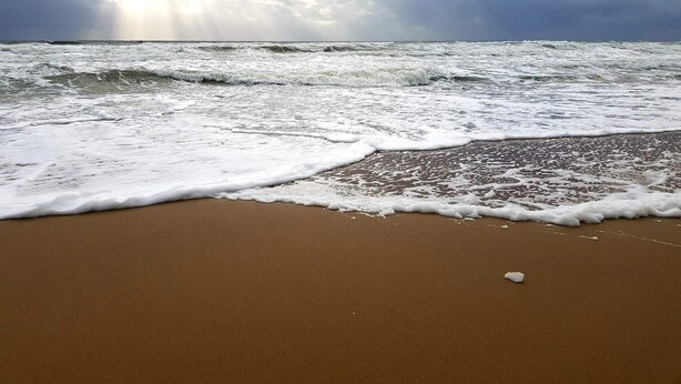 Ein Strand mit Wellen, bewölktem Himmel und sichtbarem Horizont.