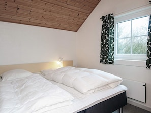 Et soveværelse med en seng, et natbord, en lampe, et vindue med gardiner og et træloft.